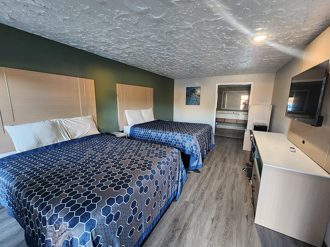A two-queen guestroom at the Americas Best Value Inn Longview hotel with blue-patterned bedspreads, desk & chair, flat-screen tv on the wall, and bathroom vanity at the back wall.