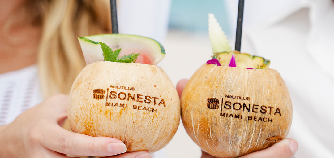Two people touching Nautilus Sonesta Miami Beach logo branded coconut drinks together.